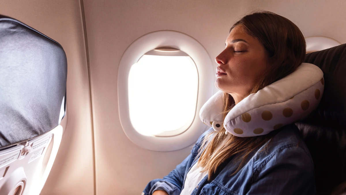 Mujer durmiendo en el avión junto a la ventanilla con una almohada de viaje en forma de U alrededor del cuello